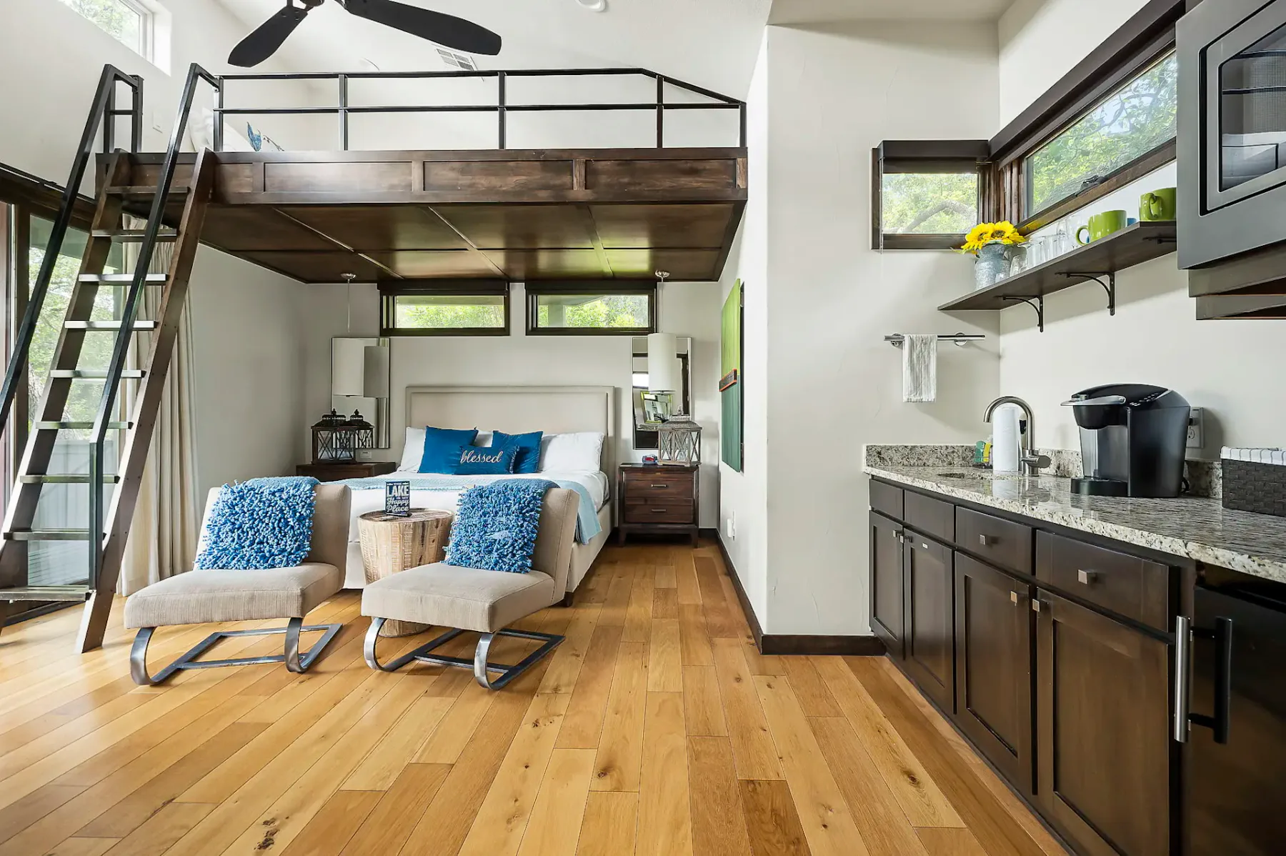 Modern studio with loft bed, wooden floors, and kitchenette. Features cozy chairs, blue accents, and large windows. Ladder leads to loft area.