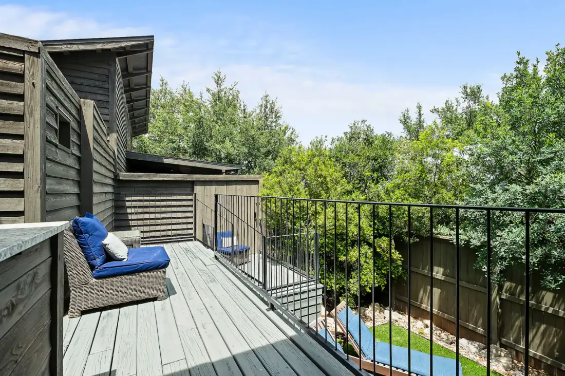 Una terraza de madera con asientos y cojines azules da a árboles frondosos.