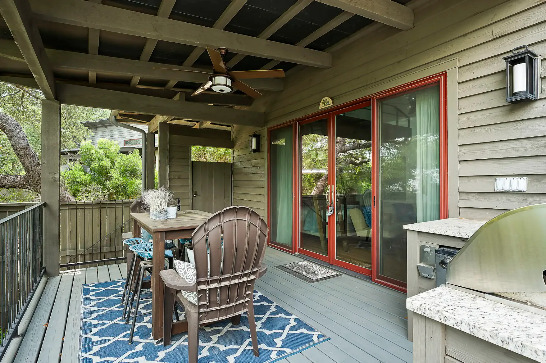 Patio cubierto con una mesa de madera, sillas, ventilador de techo y parrilla. Las puertas corredizas rojas dan al interior y la zona está rodeada de vegetación.