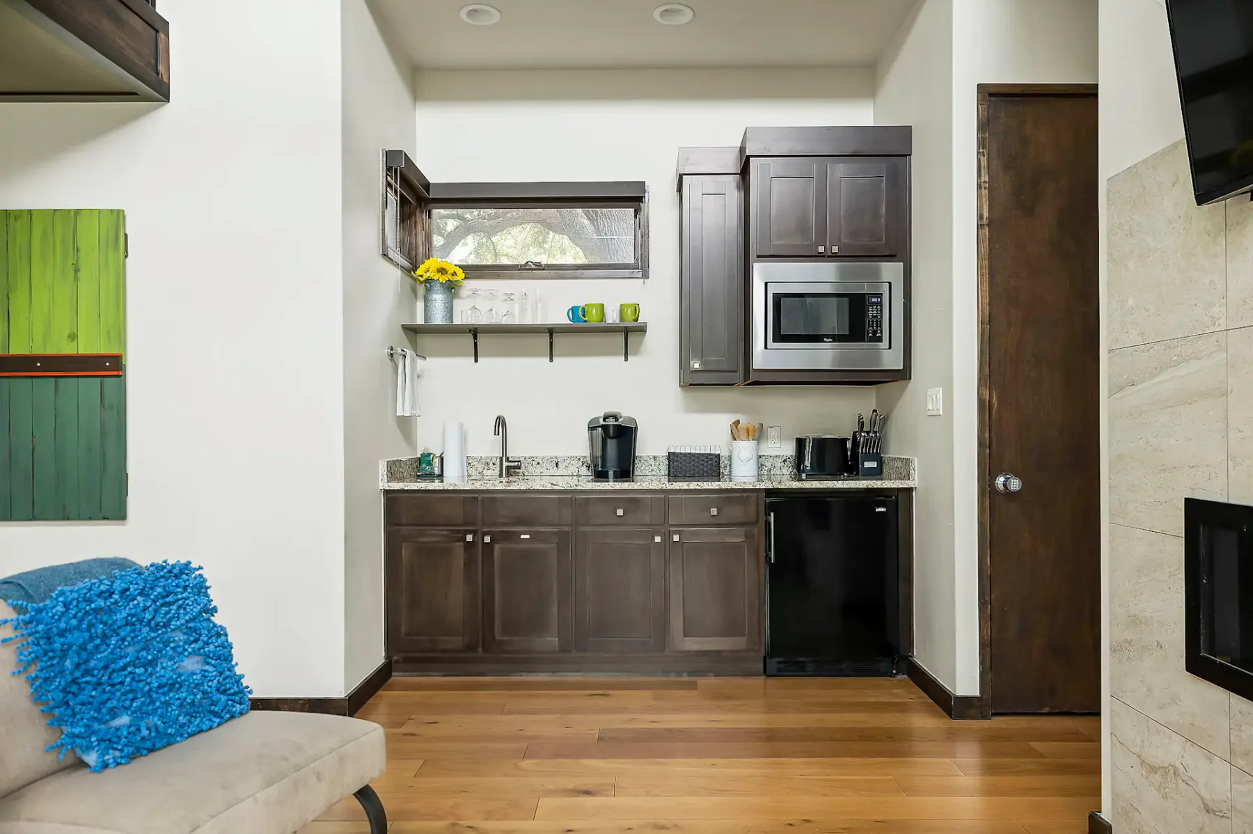 Small kitchen area with dark wood cabinets, granite countertop, microwave, and coffee maker. Bright blue pillow on nearby beige couch.