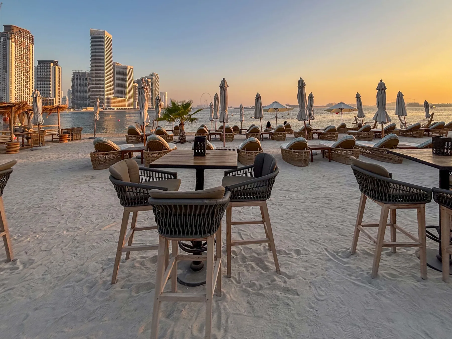 Zona de comedor al aire libre junto al agua, con mesas, asientos y sombrillas cerradas al atardecer.