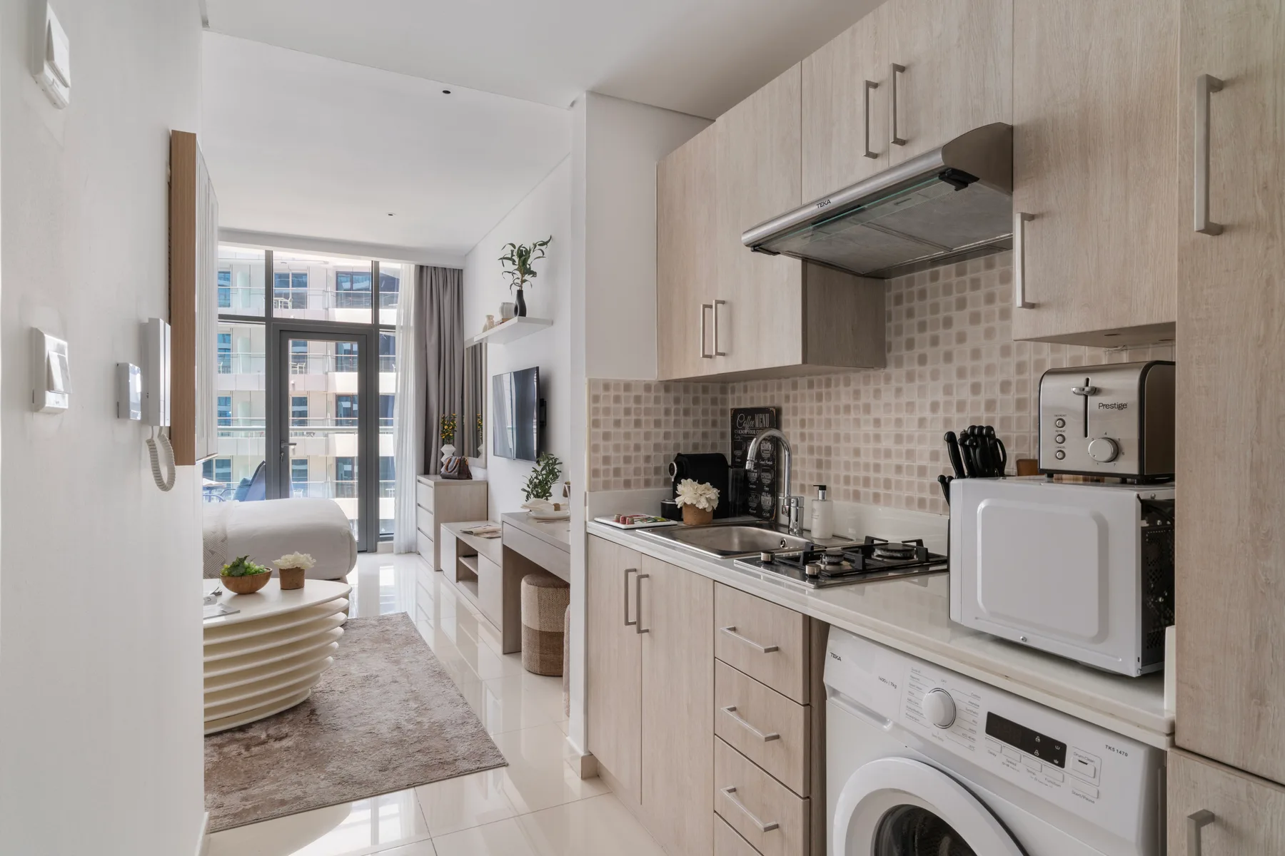 Una cocina compacta con gabinetes claros, una estufa y un microondas junto a una sala de estar.