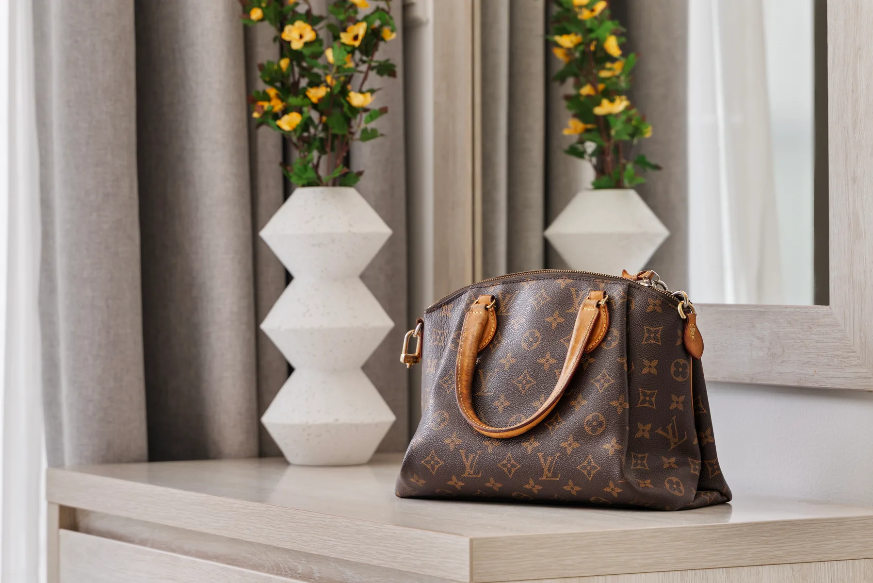 Un bolso marrón estampado está sobre una mesa clara junto a un jarrón con flores.