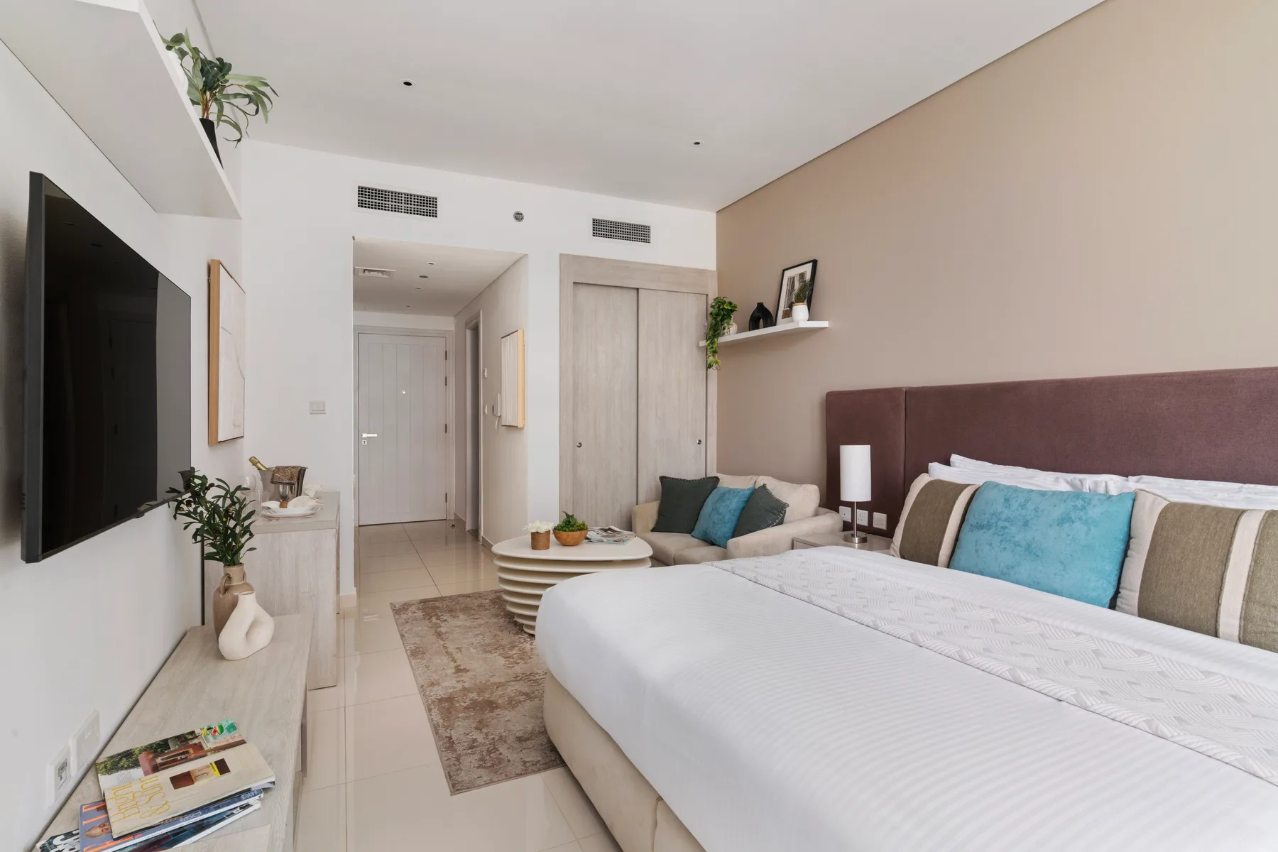 Un dormitorio luminoso con una cama grande, televisor en la pared y una pequeña zona de estar.
