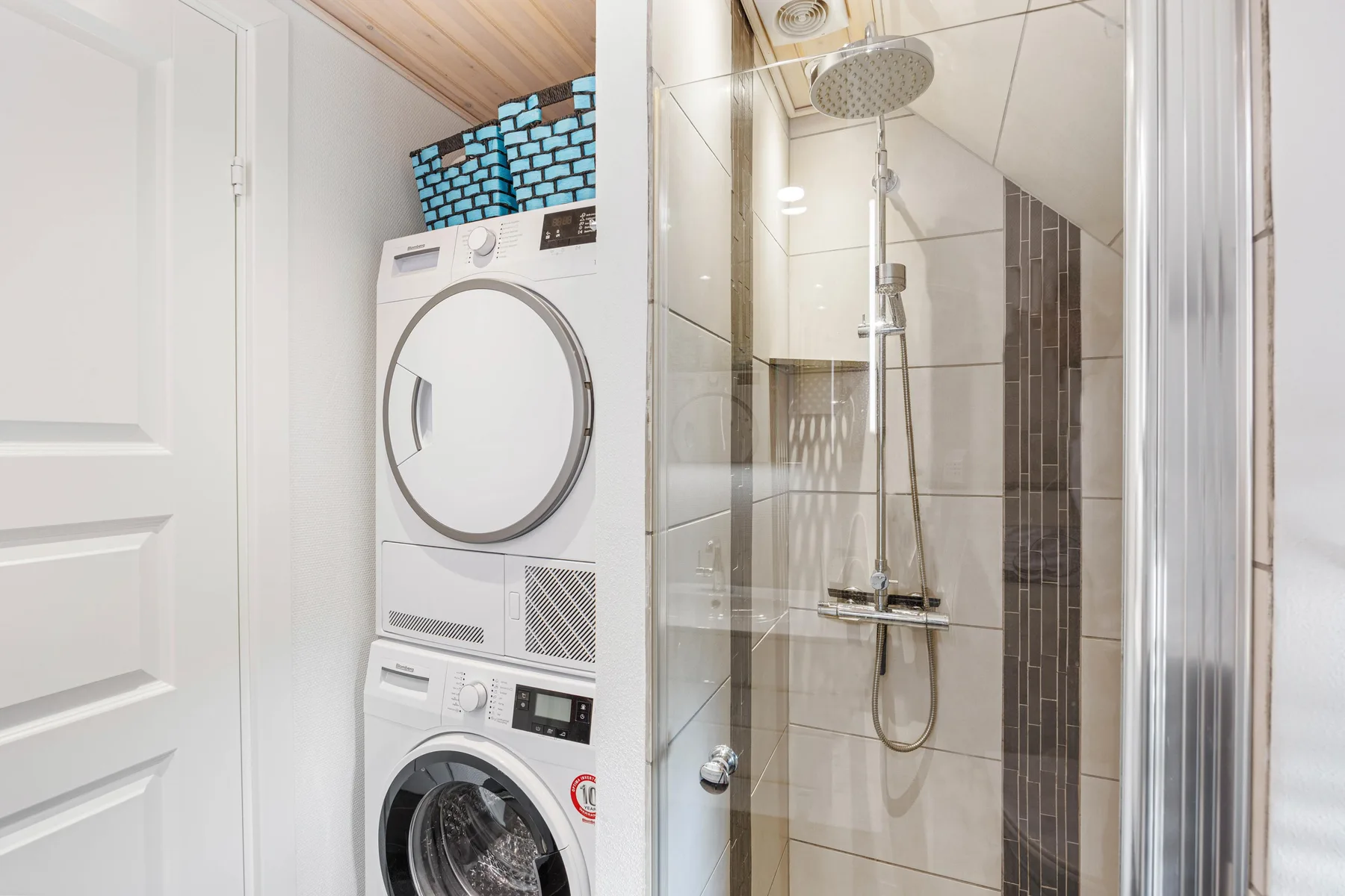 Zona de lavandería con máquinas apiladas junto a una ducha revestida de azulejos.