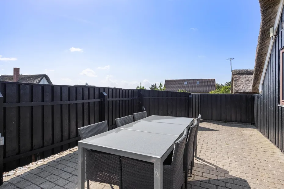 Un patio exterior tiene una mesa de comedor y asientos, rodeados por una cerca oscura.