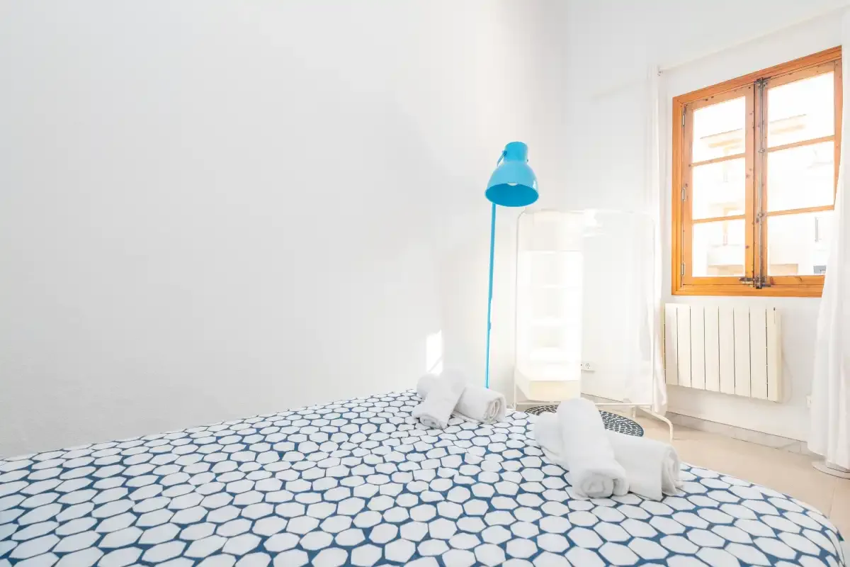 A bright bedroom with patterned bedding, a blue lamp, and a wooden-framed window.
