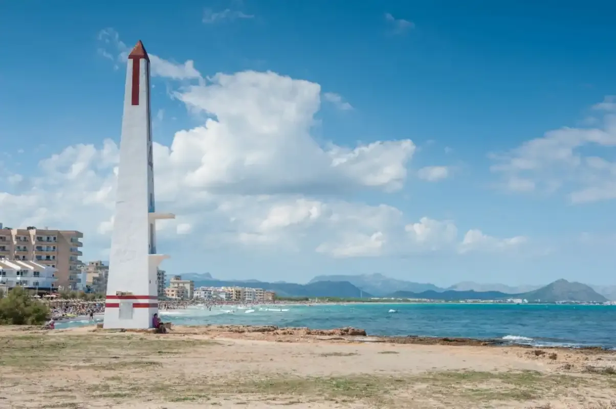 A seaside promenade beside a tall white marker, with buildings and mountains along the coast.