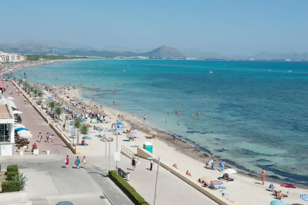 A busy beach and seaside promenade beside clear blue water.