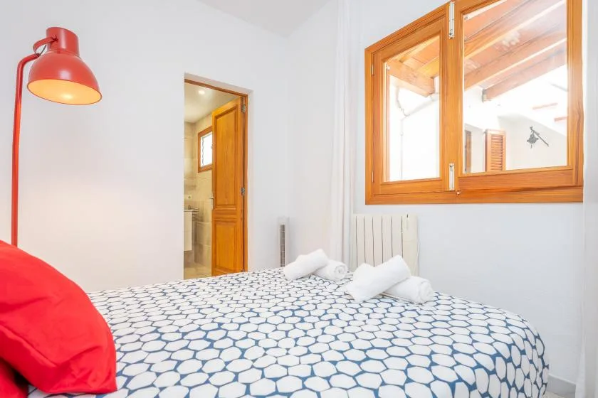 A bedroom with patterned bedding, a red reading lamp, and a wooden-framed window.