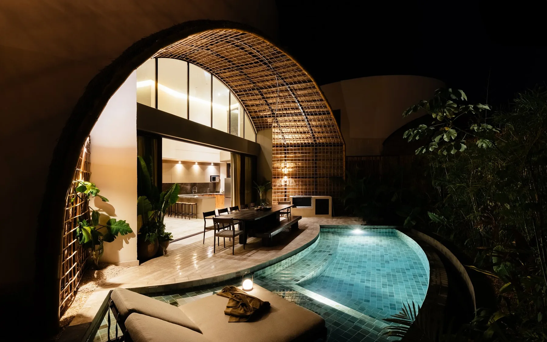 A lit patio dining area opens onto a curved pool beneath an arched canopy.