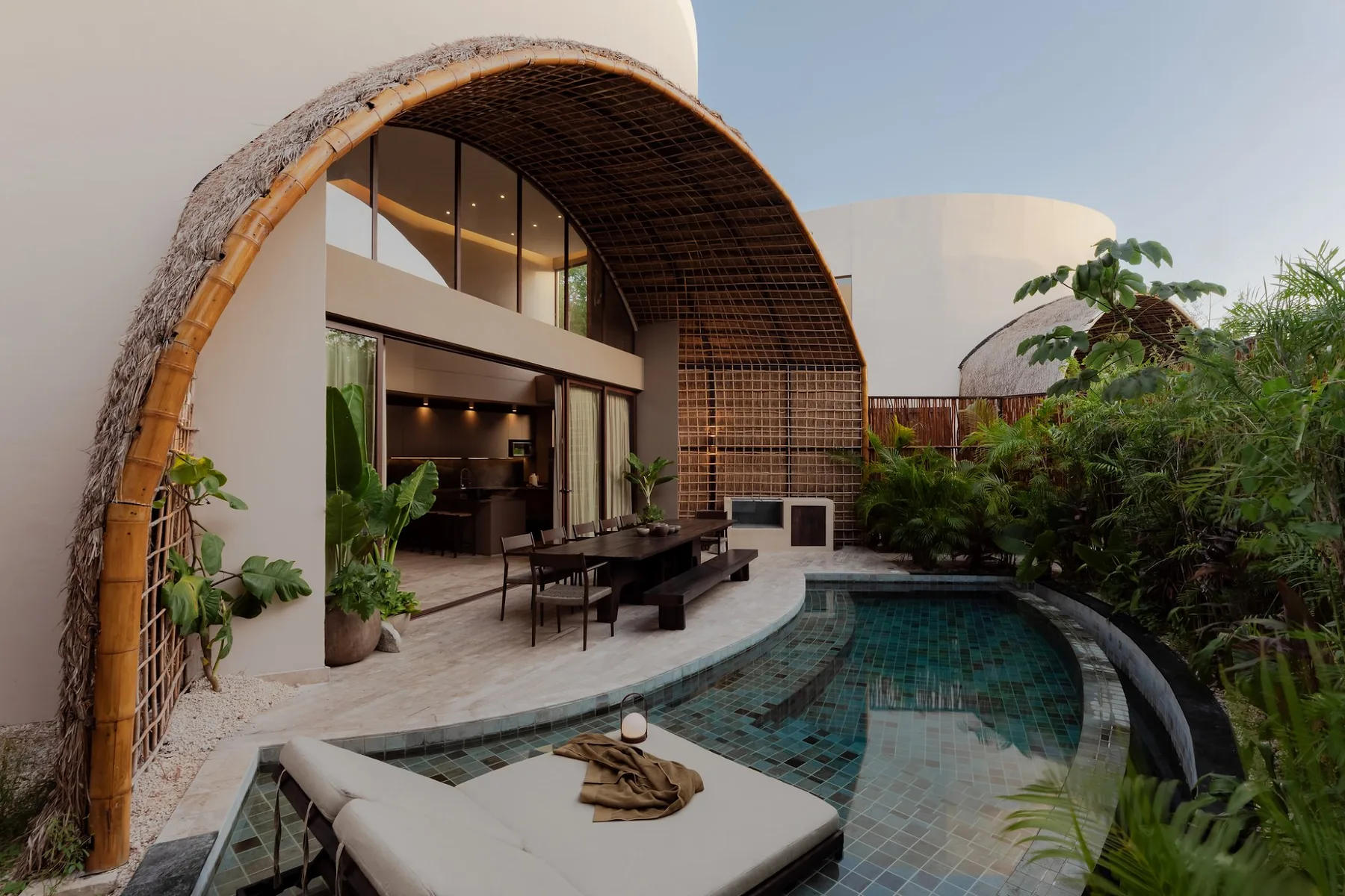 Una piscina exterior curva junto a una villa moderna, rodeada de plantas tropicales.