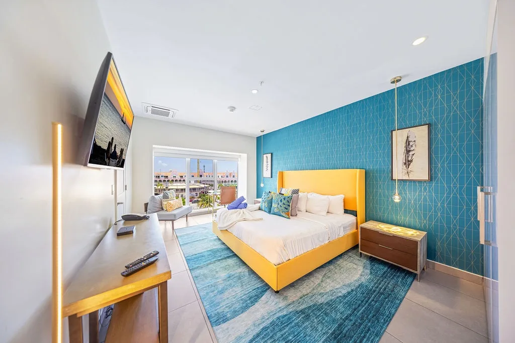 Unwind in your vibrant bedroom with a bold turquoise accent wall, cheerful yellow bed, and a balcony that lets in warm sunshine—your stylish coastal retreat awaits.