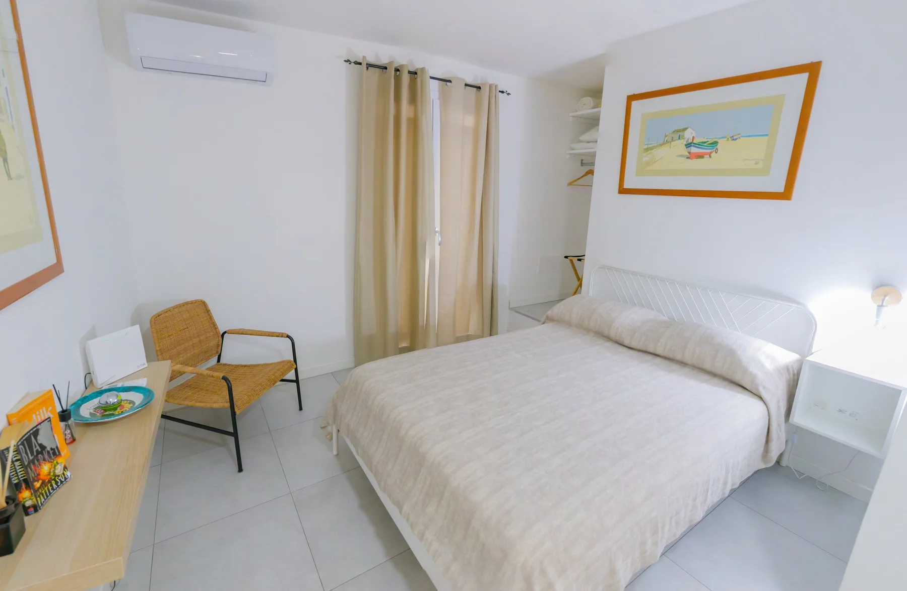 Un dormitorio luminoso con una cama tendida, cortinas y una pequeña zona para sentarse.