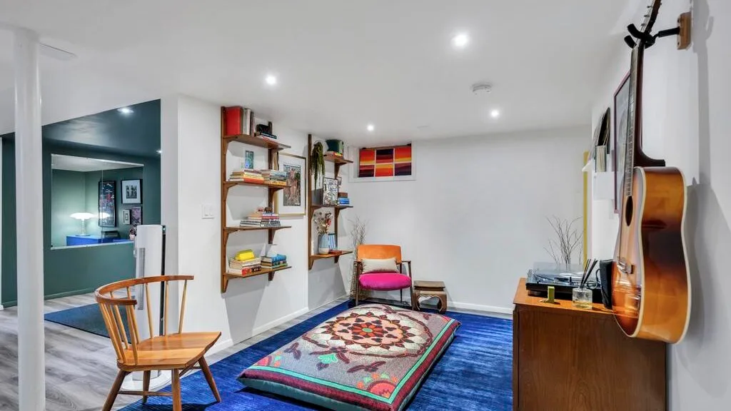 Una habitación amueblada tiene una alfombra estampada, asientos de madera, estantes y una guitarra.