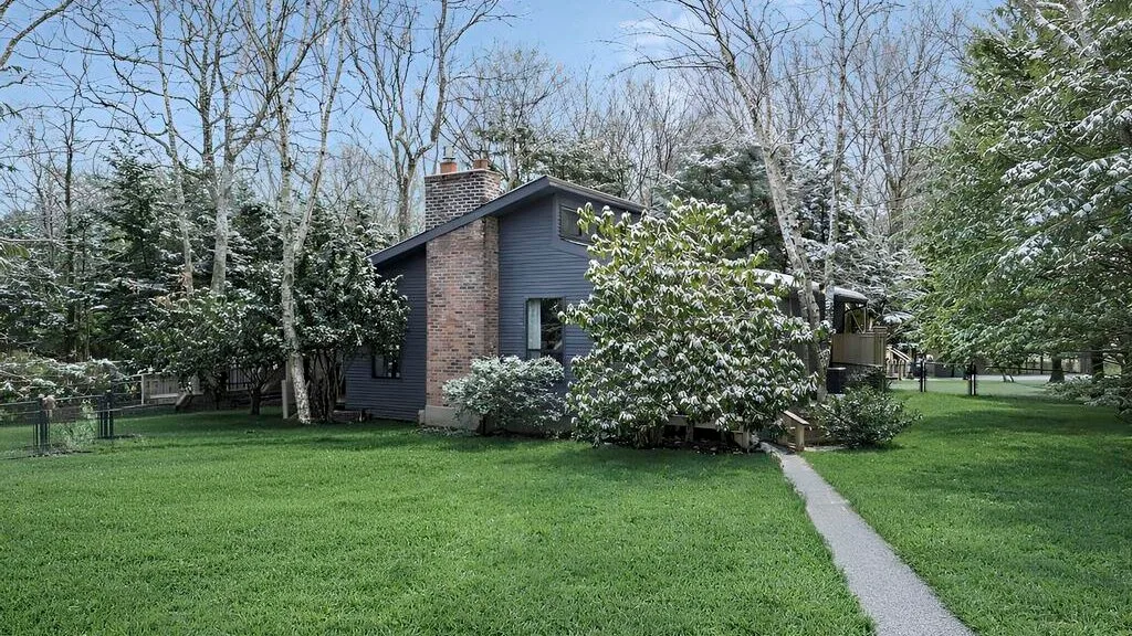 Una casa está junto a un sendero pavimentado en un césped verde, rodeada de árboles y arbustos.