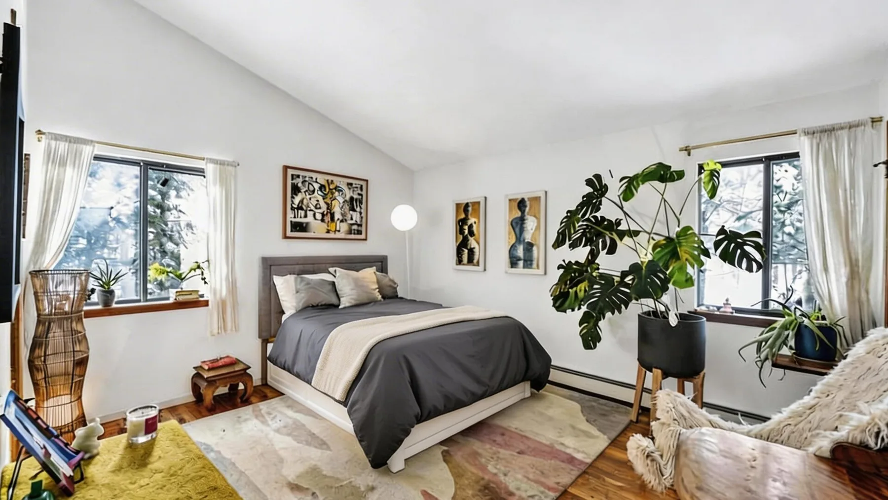 Un dormitorio luminoso con una cama, cuadros enmarcados y plantas frondosas junto a las ventanas.