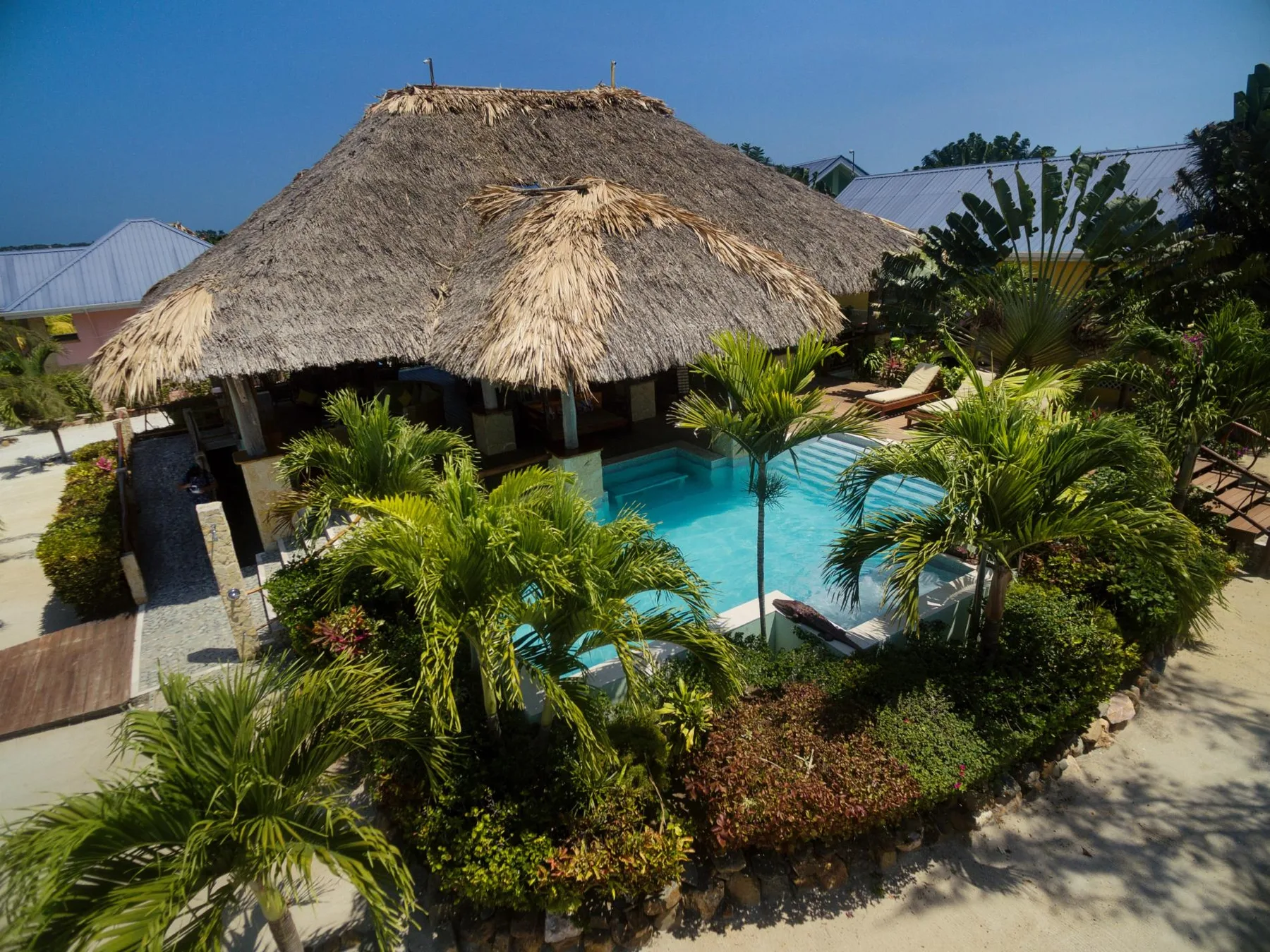 A swimming pool surrounded by tropical plants sits beside a thatched-roof pavilion.