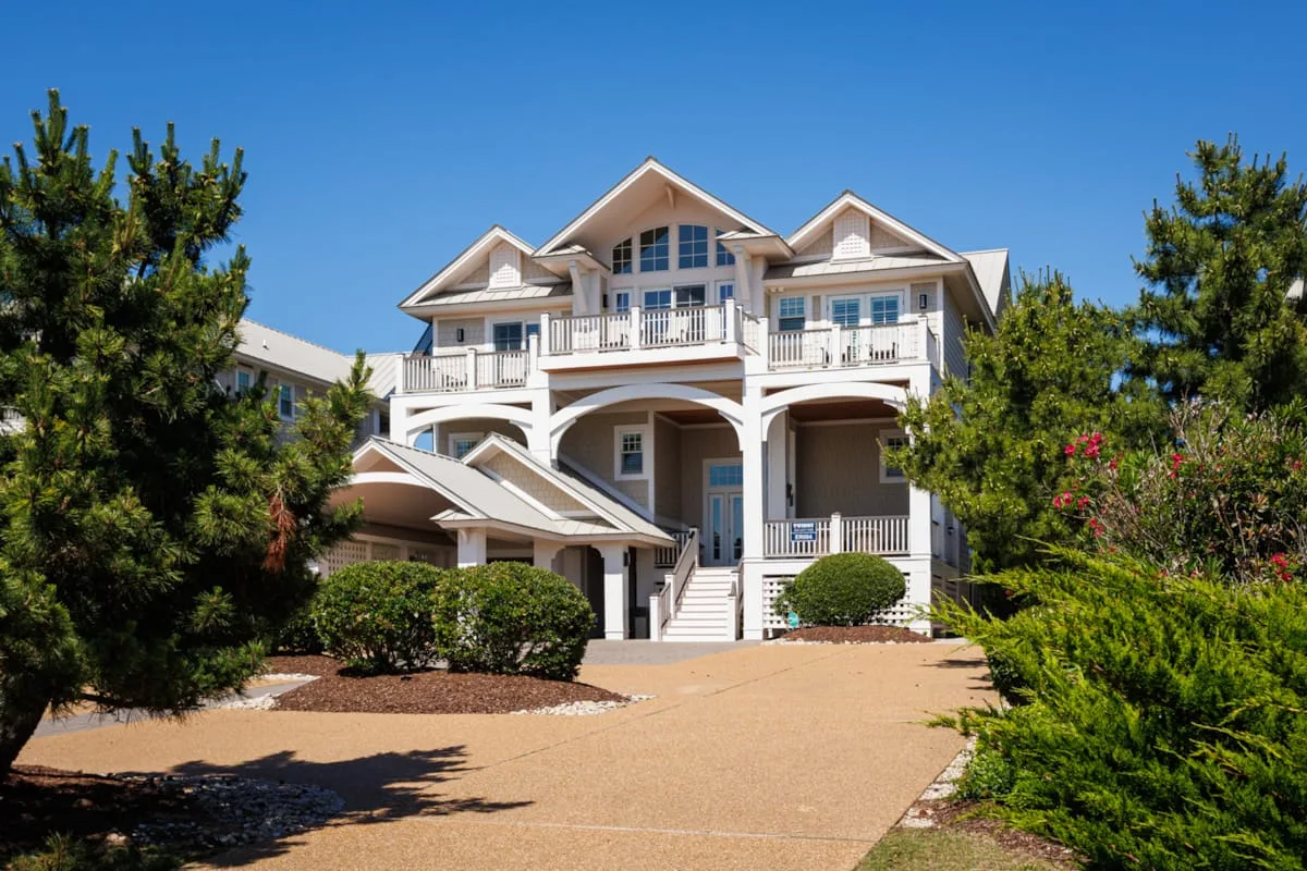 A large white house stands at the end of a paved driveway, surrounded by trees and shrubs.
