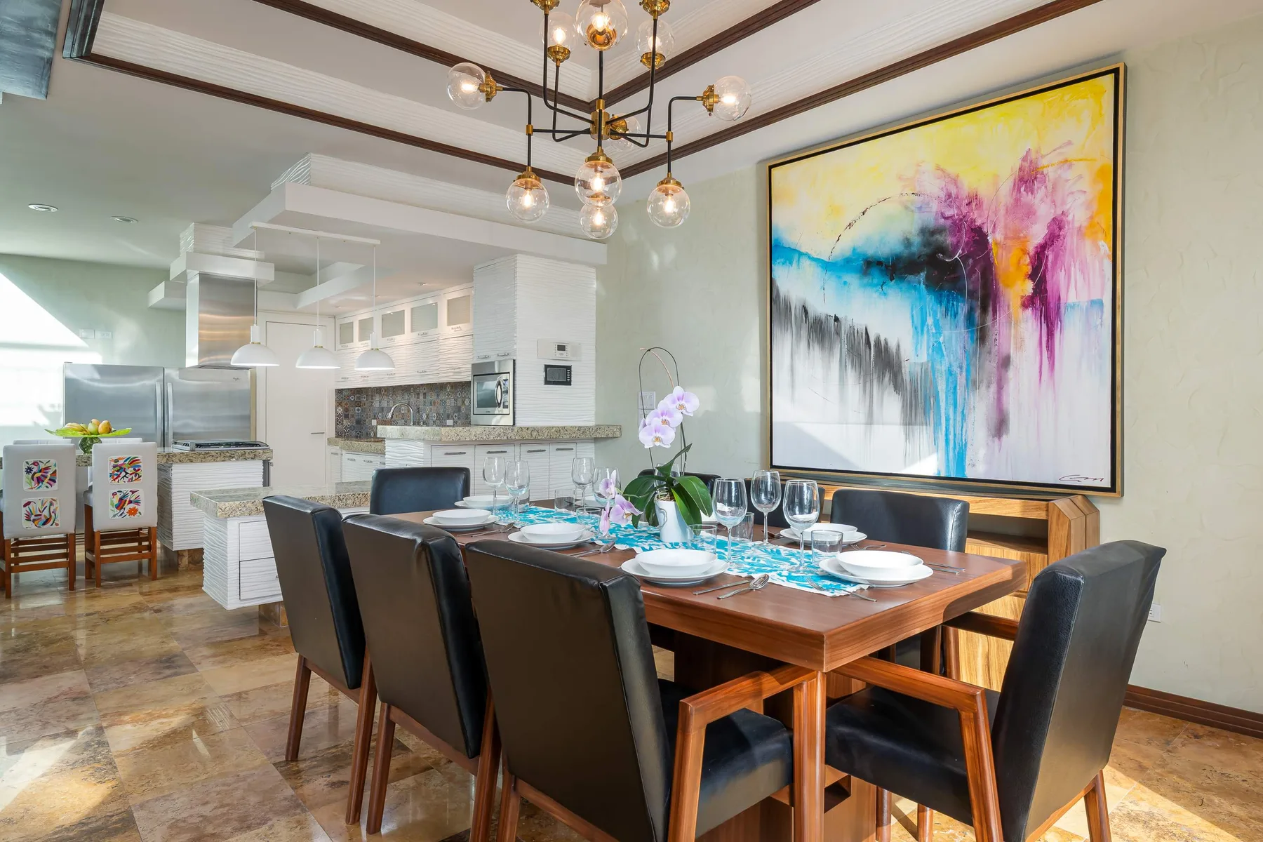 A dining area features a wooden table set with plates and dark seating beneath colorful abstract artwork.