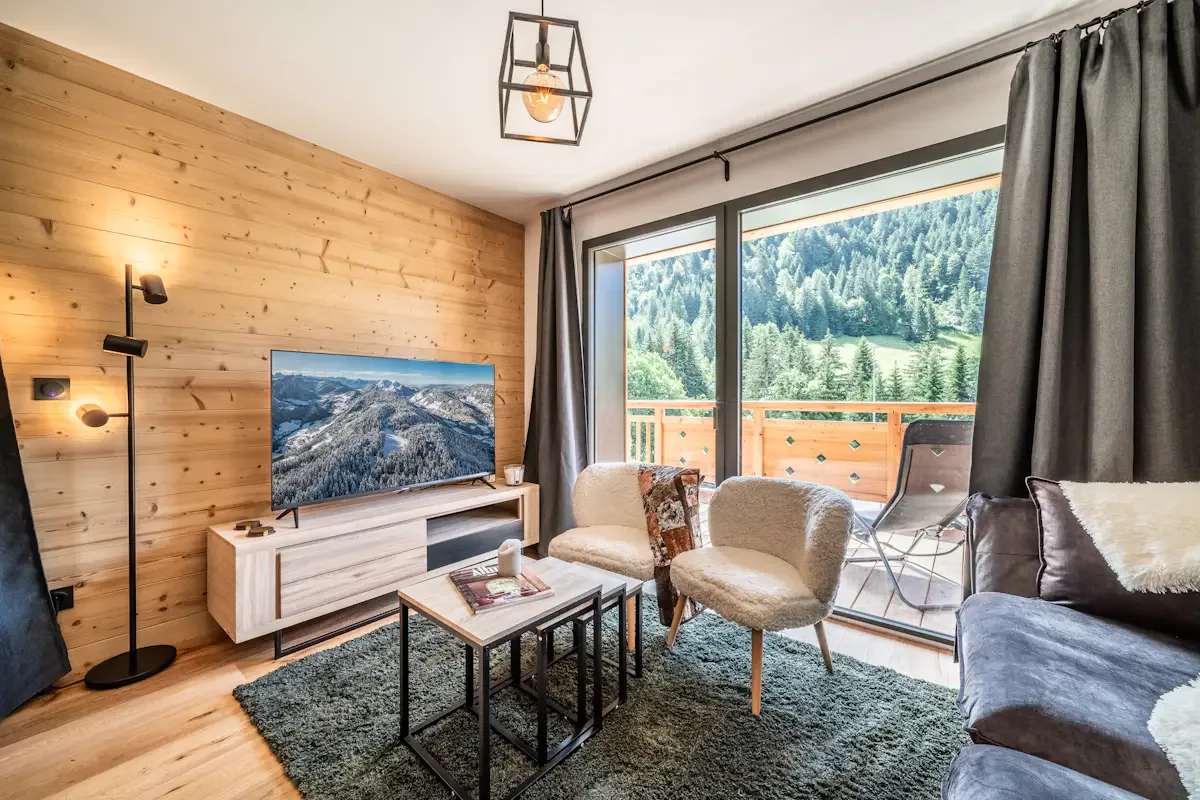 Una sala de estar con asientos, un televisor y grandes ventanales con vistas a montañas boscosas.