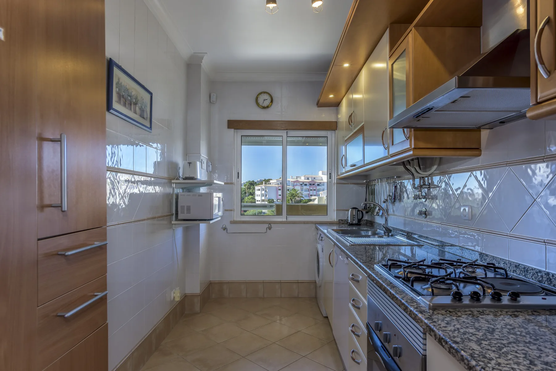 A narrow kitchen with wooden cabinets, a countertop, and a window overlooking buildings.