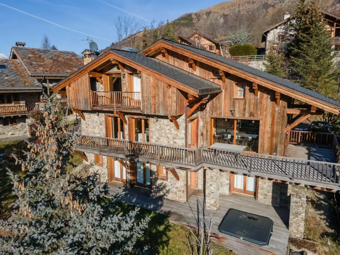 A large wooden chalet with balconies and a stone lower facade sits on a hillside.