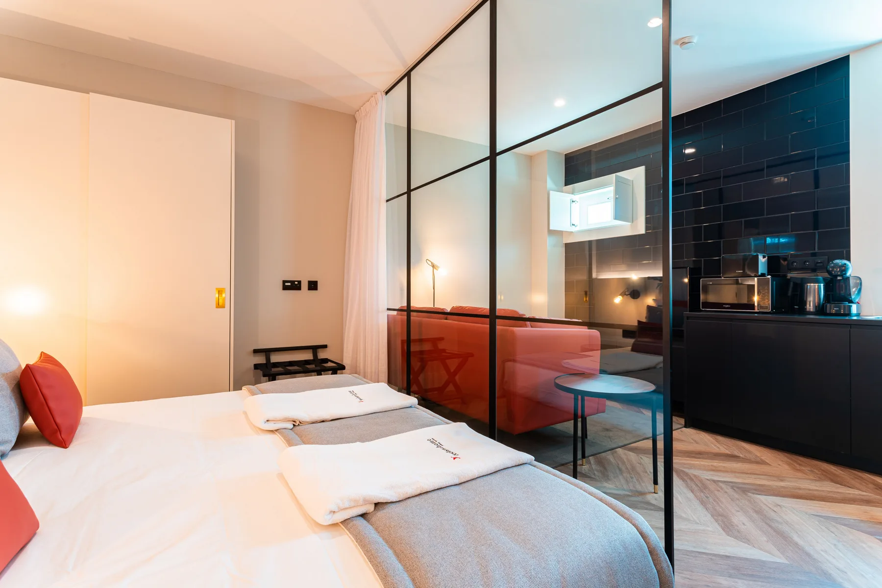 A bedroom with white bedding and a glass partition beside the kitchen.