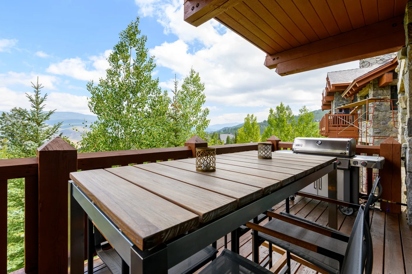 Una terraza exterior de madera con una mesa y una parrilla da a un paisaje arbolado.