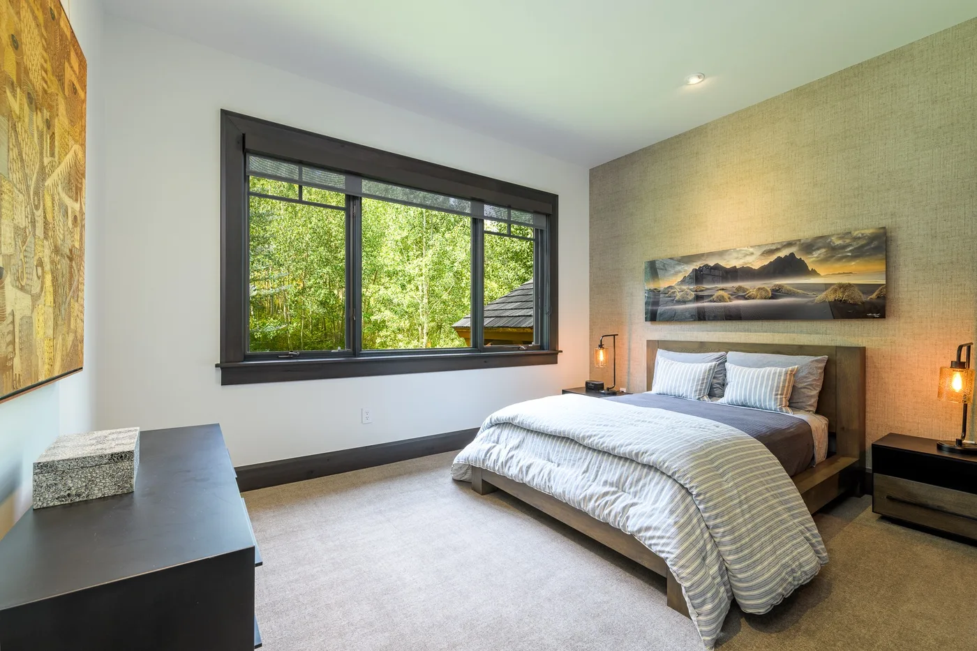 Dormitorio con una cama bajo una amplia ventana con vistas a la vegetación.
