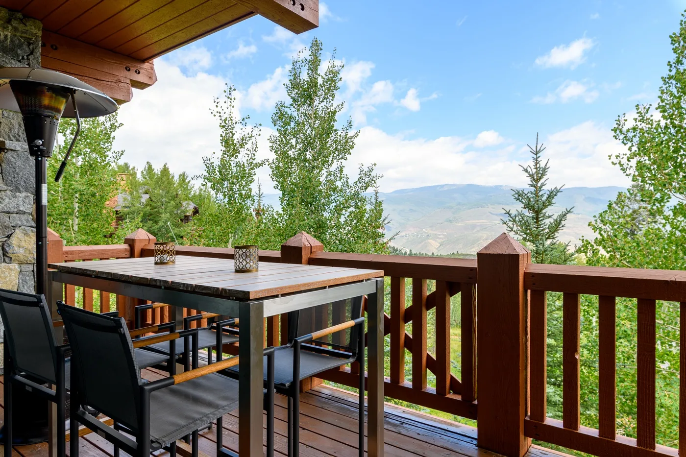 A wooden outdoor deck has a dining table and chairs overlooking a green mountain landscape.