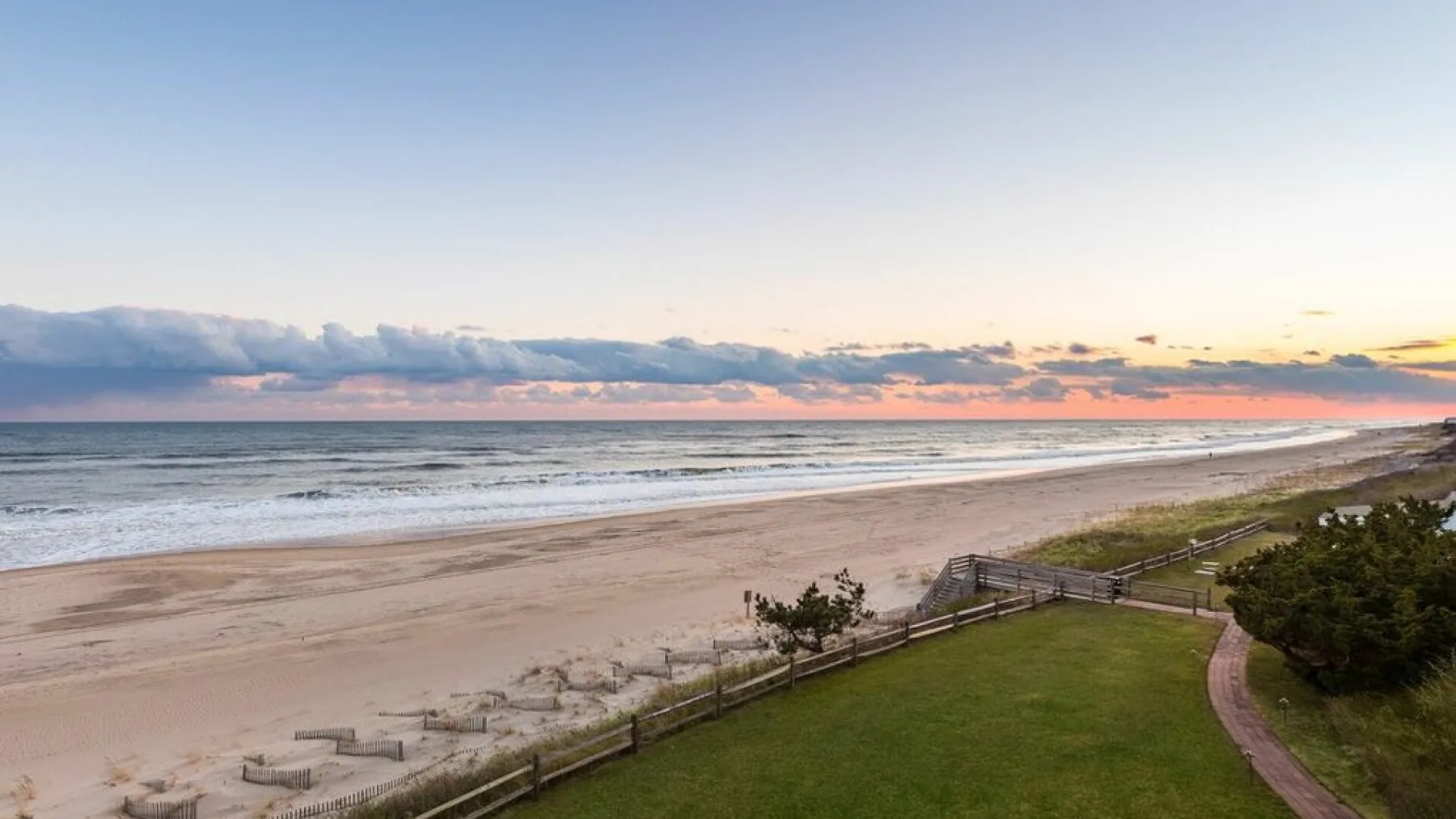 A beach and ocean stretch beside a green lawn under a colorful sunset sky.