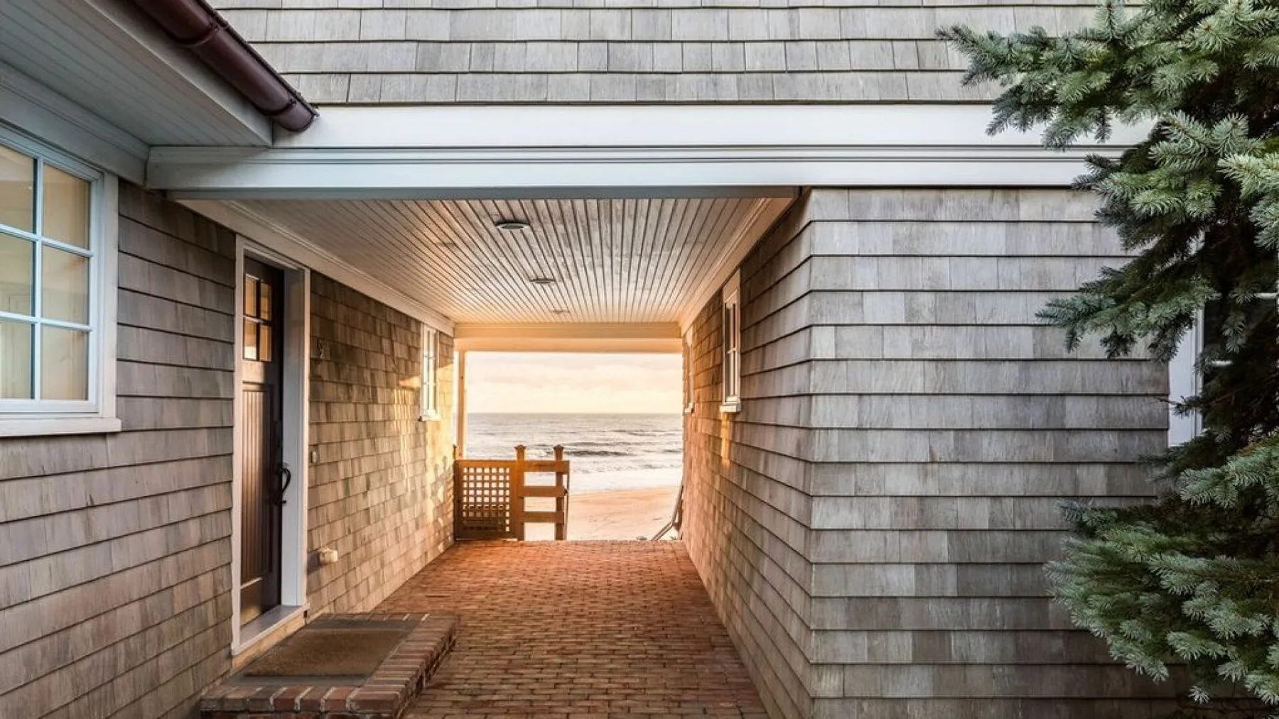 A covered passage between shingled walls opens toward a beach and the sea.