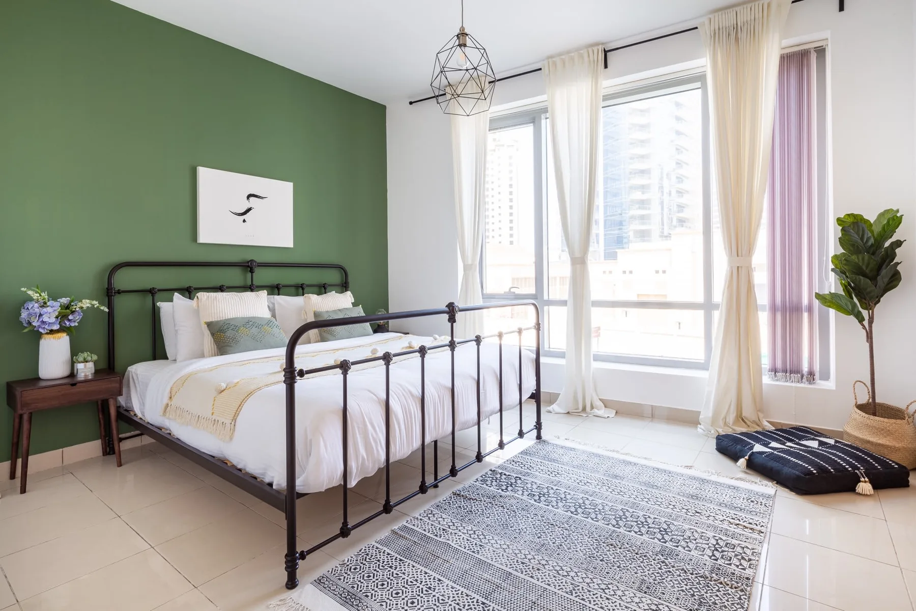 Un dormitorio luminoso con cama de estructura metálica, pared verde y grandes ventanas con cortinas.