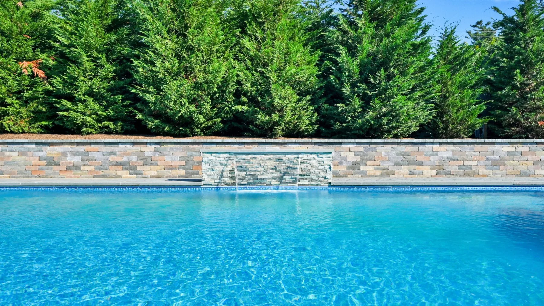 Una piscina al aire libre está junto a un muro de piedra y árboles altos.