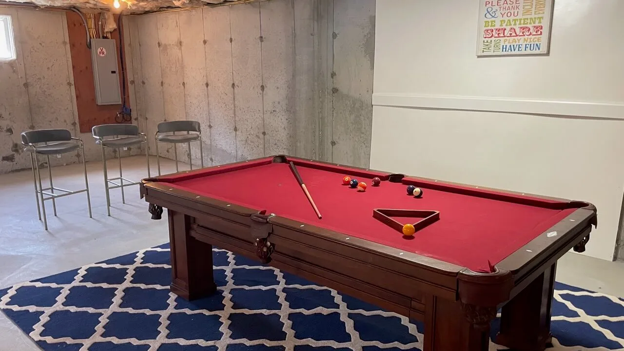 Una mesa de billar con paño rojo está en una sala de recreo del sótano, con asientos cerca.