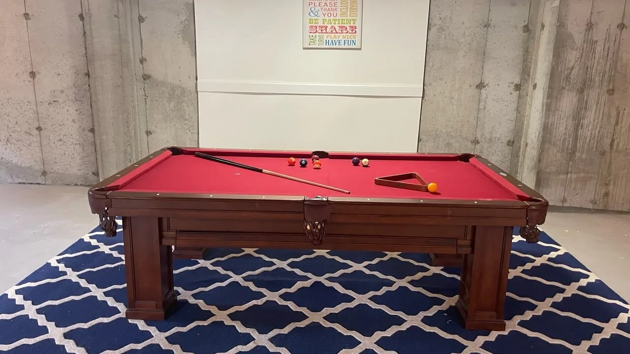 Una mesa de billar con paño rojo está sobre una alfombra azul estampada en una sala con paredes de concreto.