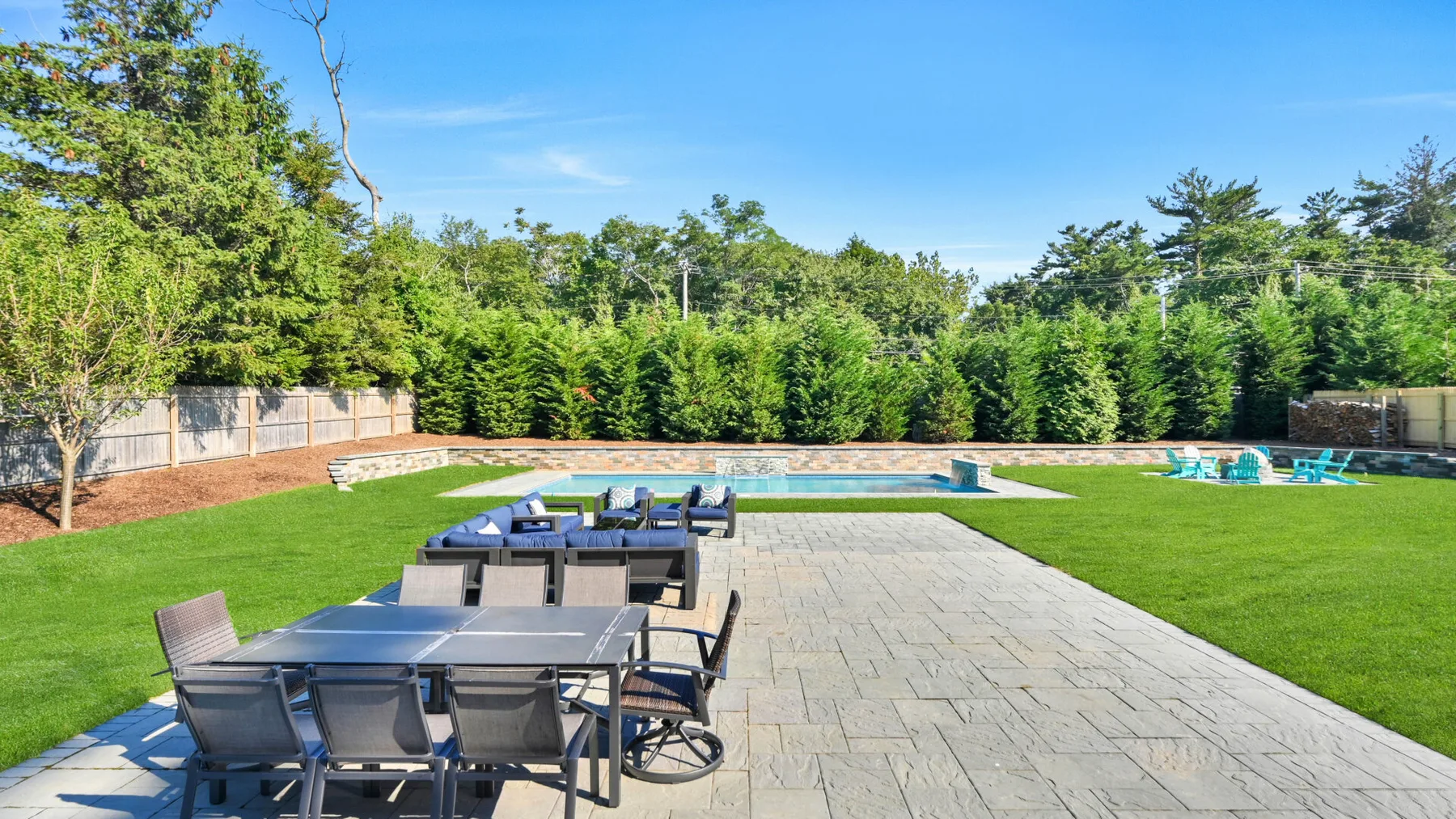 Un patio pavimentado con asientos al aire libre da a una piscina y un césped verde.