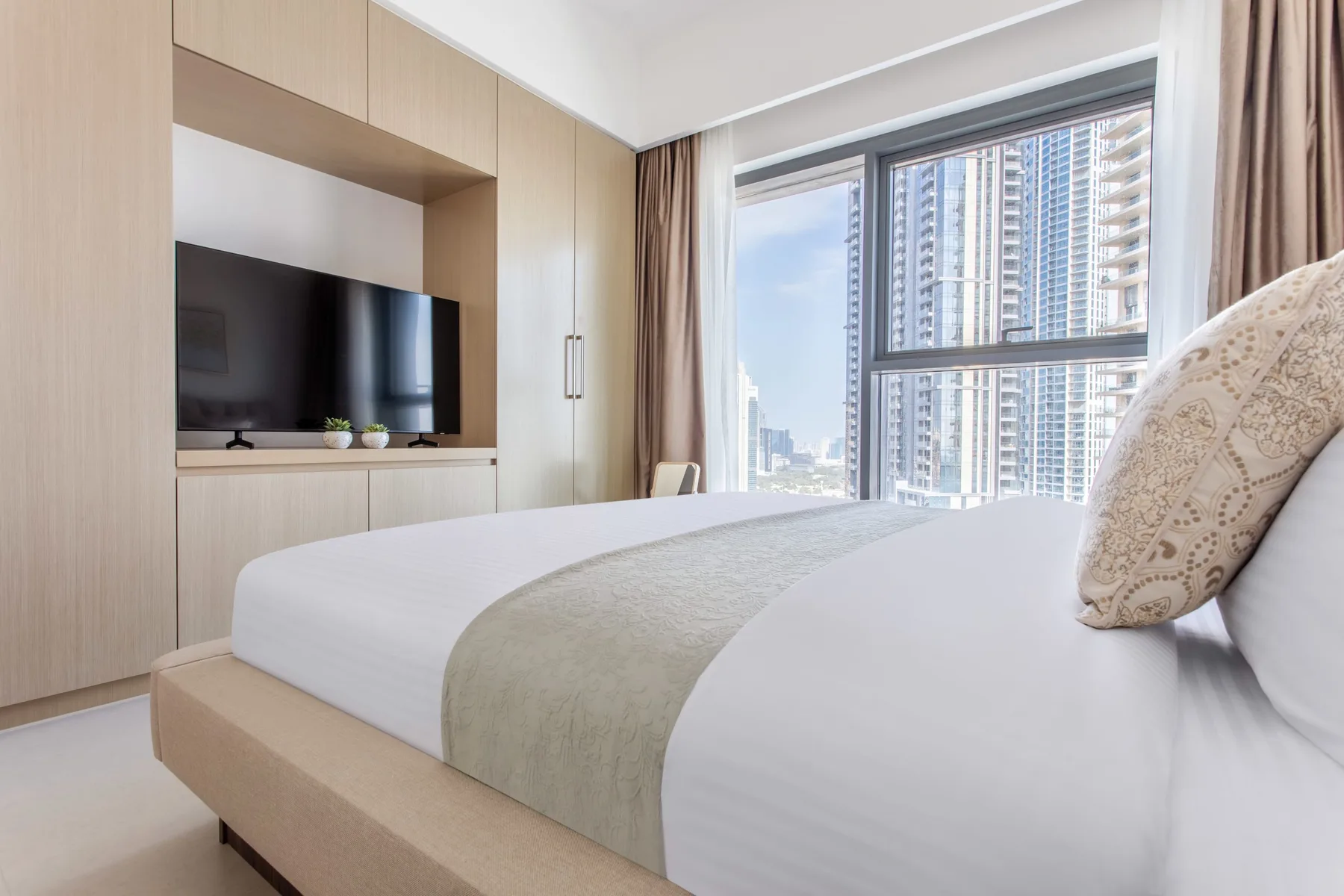 Un dormitorio luminoso con una cama bien tendida y una gran ventana con vista a edificios cercanos.
