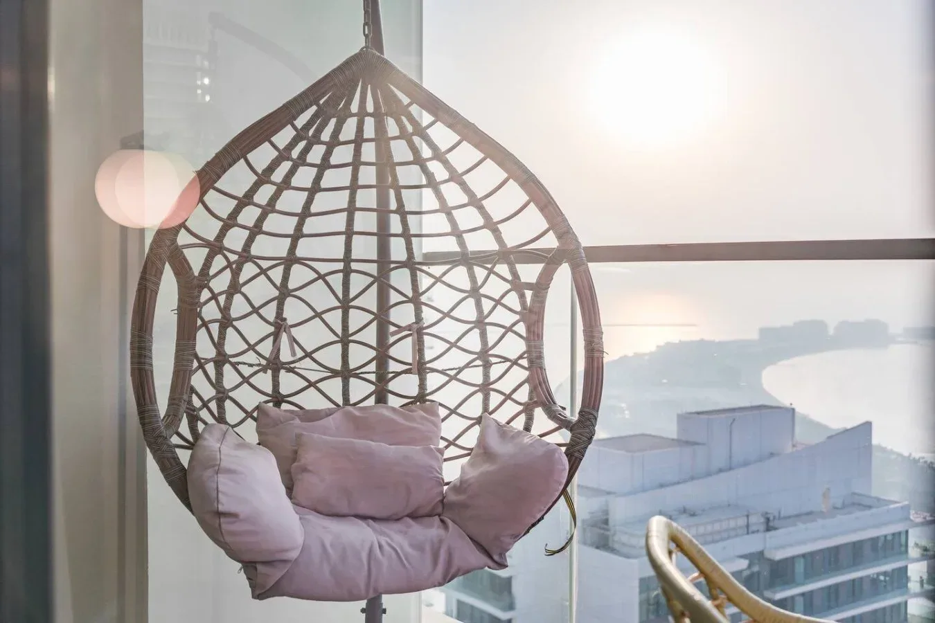 Una silla colgante tipo cesta está junto a una ventana luminosa con edificios a la vista.