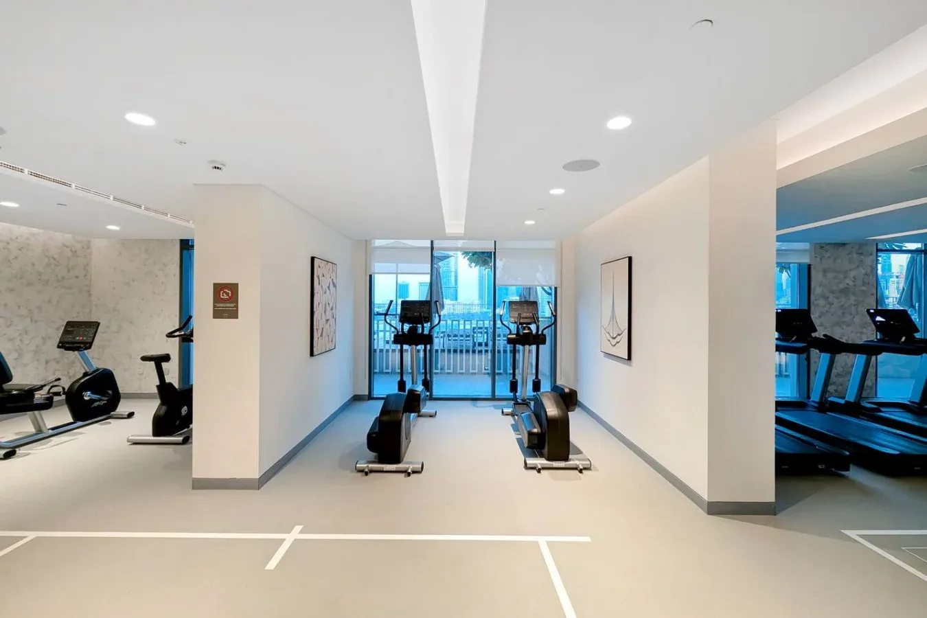 Un gimnasio interior luminoso con máquinas de ejercicio y grandes ventanales al fondo.