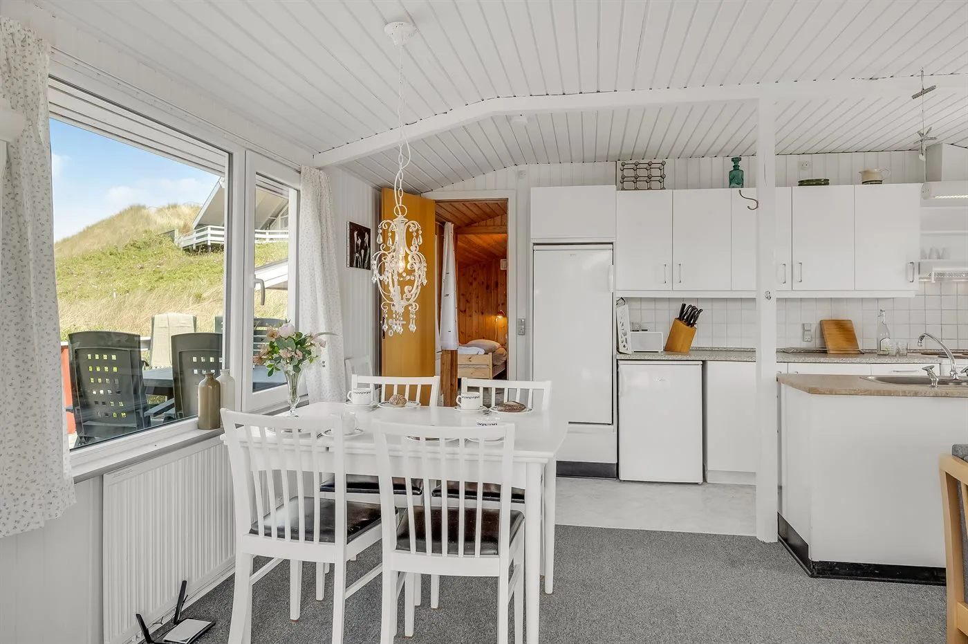 Un comedor luminoso se abre a una cocina blanca con grandes ventanas.