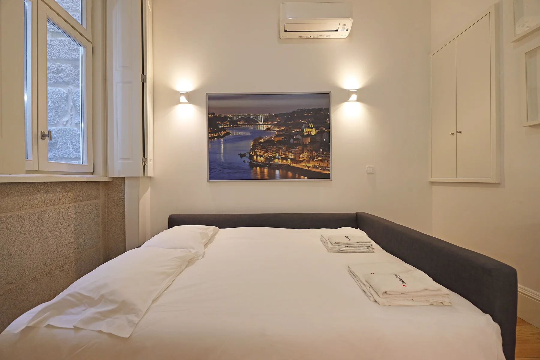 A bedroom with a neatly made bed and a framed cityscape above it.
