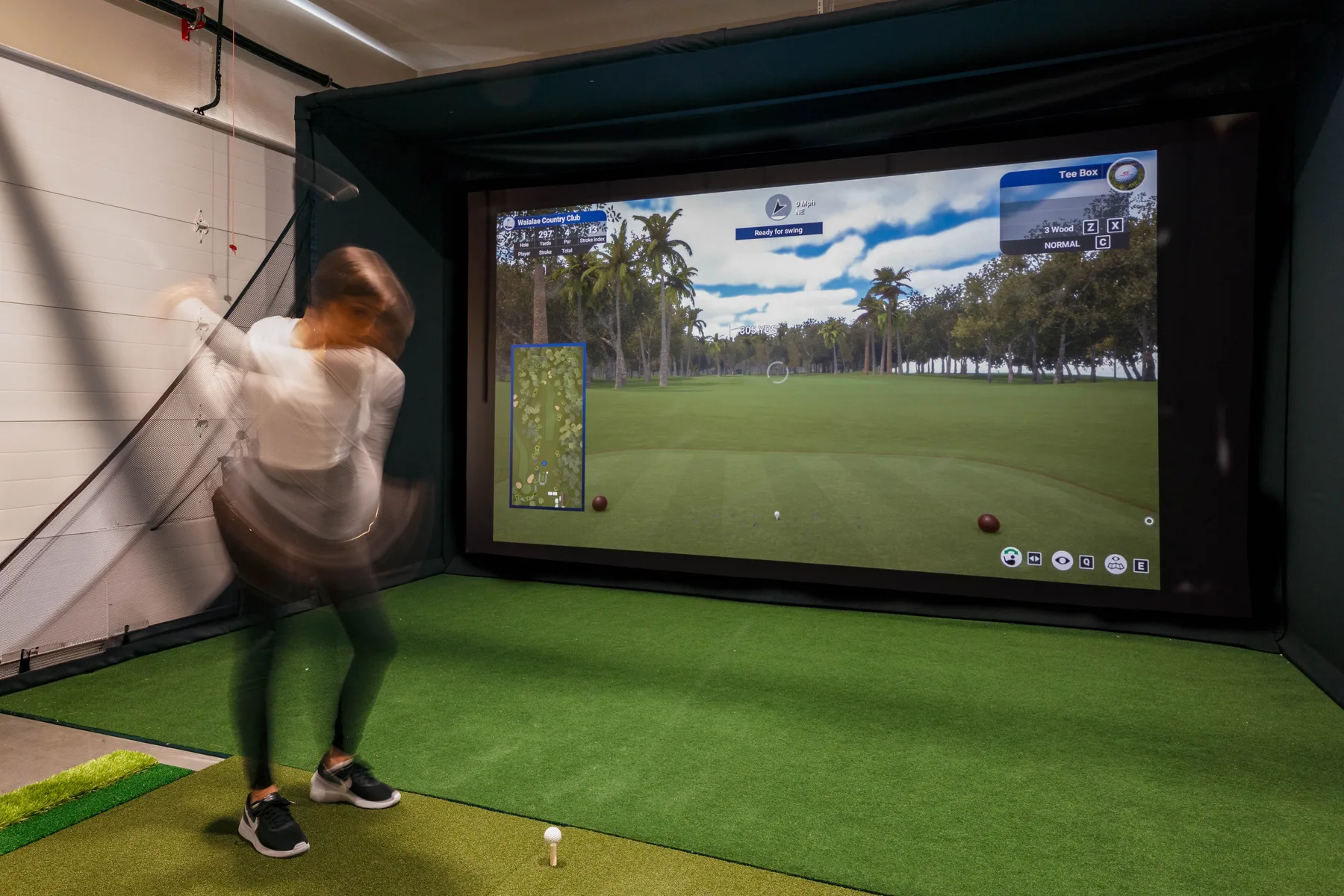 An indoor golf simulator displays a virtual course beside a person swinging a club.