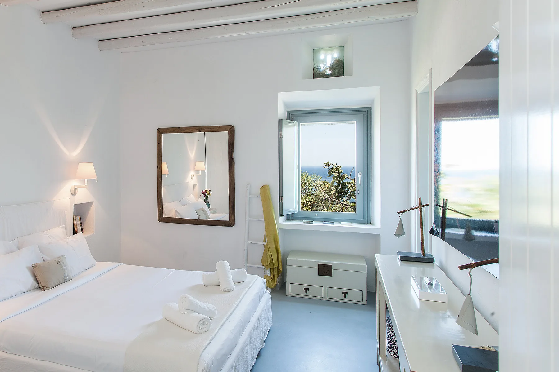 Un dormitorio luminoso con una cama, lámparas de noche y una ventana con vistas al mar.