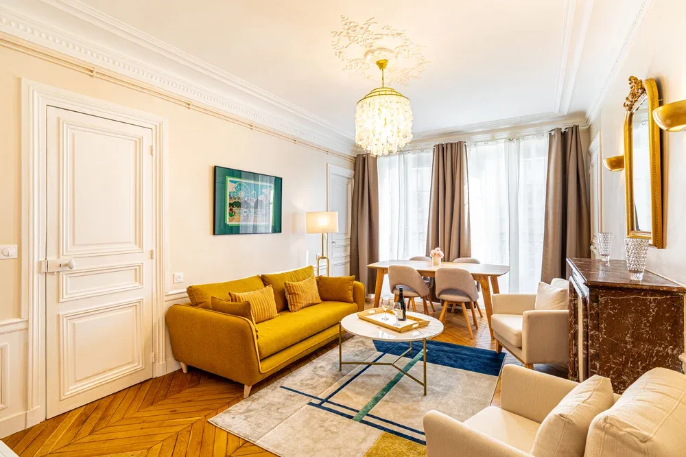 A bright living room with mustard-colored seating, a coffee table, and tall curtained windows.