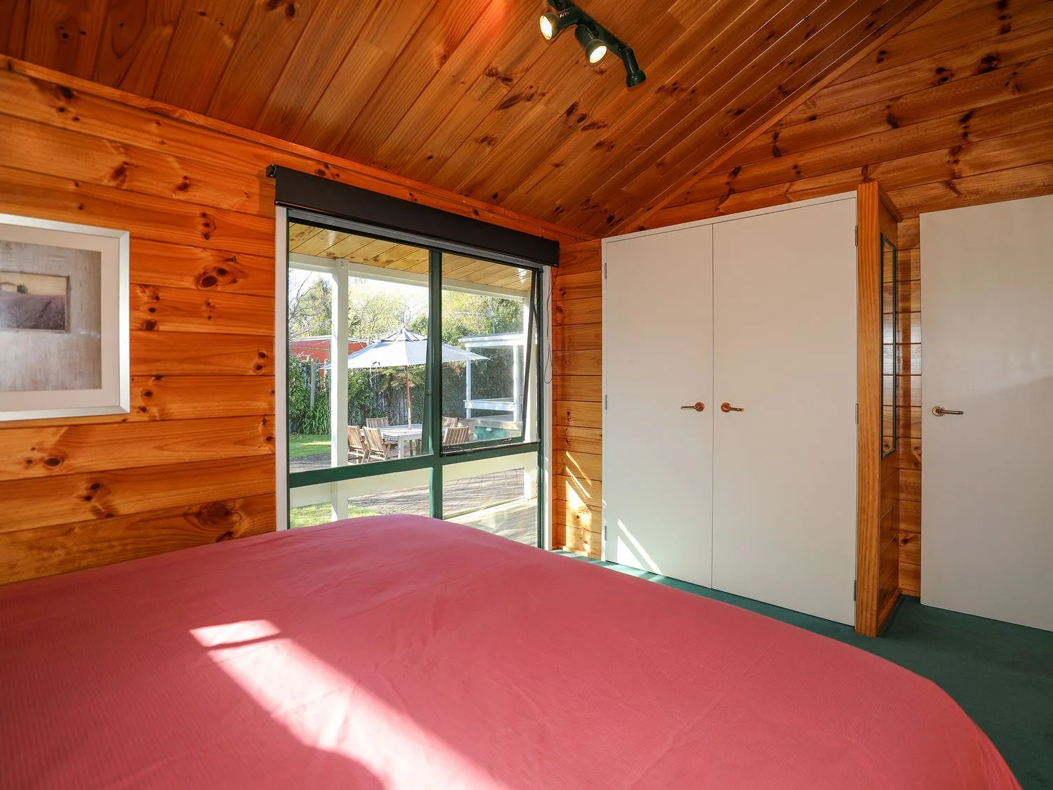 Un dormitorio con una cama cubierta con una manta roja, paredes de madera, una ventana que da a una zona de asientos al aire libre y puertas dobles blancas en Five Mile Yay - Five Mile Bay Holiday Home, en Waitahanui, a 12 km de Taupo.
