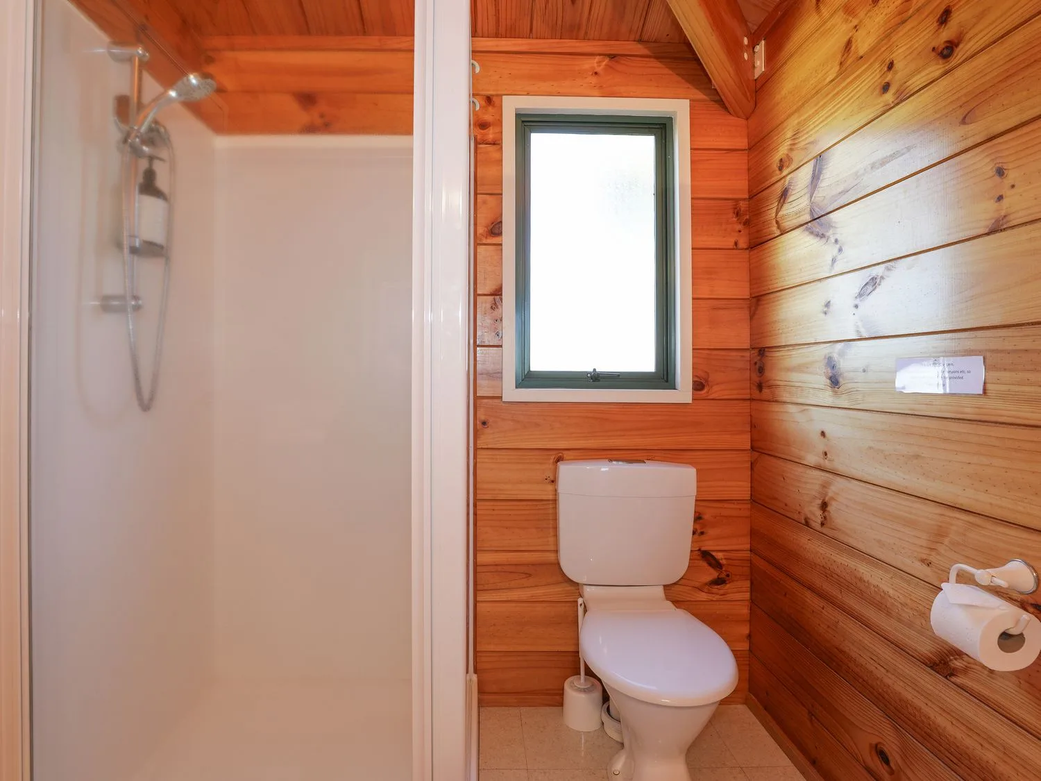 Un baño con ducha, inodoro y ventana, con paredes revestidas de madera.