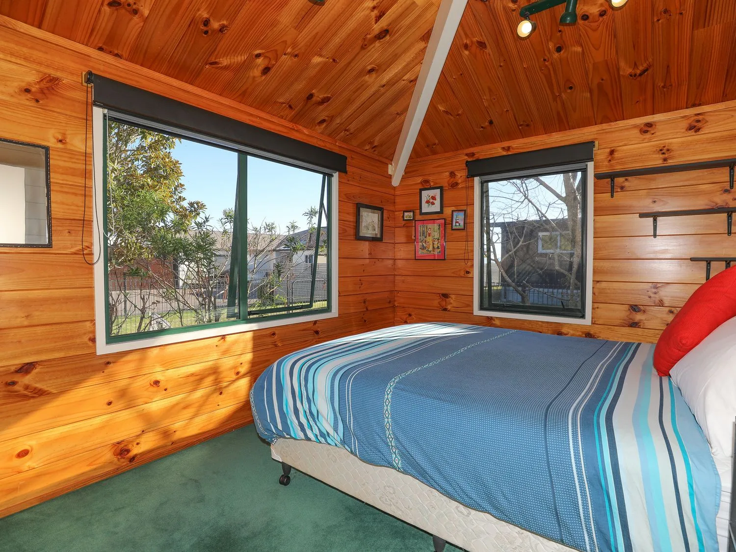 Un dormitorio con paredes y techo de madera, dos ventanas y una cama con ropa de cama de rayas azules y una almohada roja en Five Mile Yay - Five Mile Bay Holiday Home, en Waitahanui, a 12 km de Taupo.