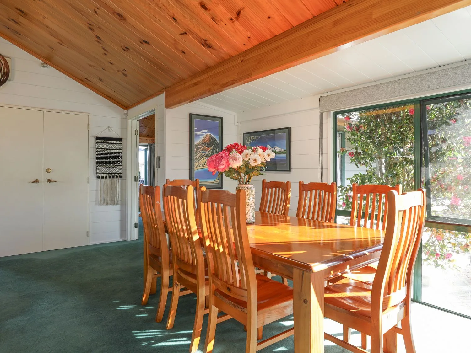 Un comedor con una mesa de madera, asientos, flores y ventanas grandes.